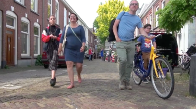 A family walks four abreast down a brick-paved street. A man wearing a blue shirt in the center walks a yellow bicycle