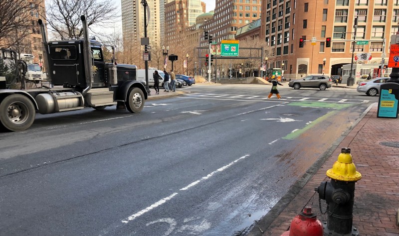 Atlantic Avenue at its intersection with Oliver Street and Seaport Boulevard in downtown Boston, showing pedestrians crossing the street in front of a large truck and the I-93 on-ramp in the distance.
