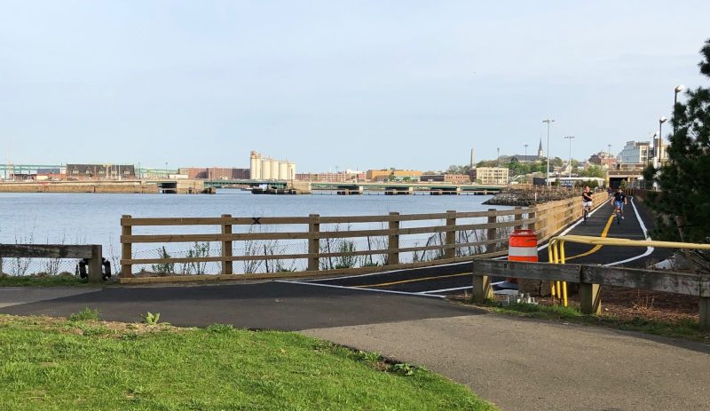 The northern entrance to the new Charlestown Seawall bike path, pictured from Draw 7 Park in Somerville. The Alford Street Bridge and the spire of the Saint Vincent de Sales Church on Bunker Hill are visible in the distance.