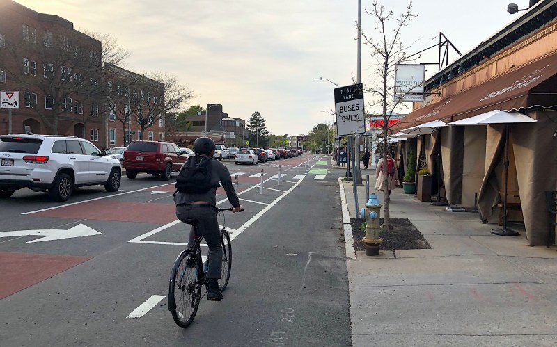A bike rider rides along a flexpost-protected bike lane next to a sidewalk cafe on one side and a line of cars waiting in traffic on the other..