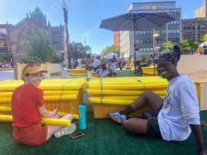 Dos estudiantes de arquitectura, Miranda y Denzel, encadenando tubos de espuma para la piscina sobre cajas de madera - una de las varias opciones disponibles a la gente para sentarse durante Copley Connect, un proyecto piloto que cerrará una cuadra de la calle Dartmouth a vehículos para poder ampliar el espacio público de Copley Square.