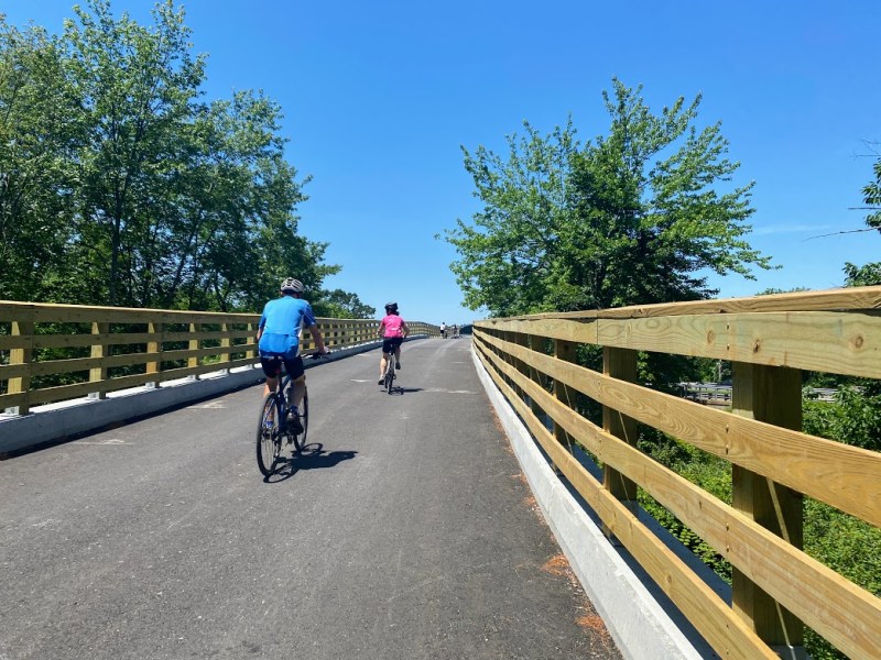 Although the Bruce Freeman Rail Trail bridge over Route 2 is still officially closed for construction, people are making their way around the existing barricades and riding their bikes on it. The left photo shows the paved portion on the south side of Route 2, while the right photo shows the unfinished, gravel portion on the northside just a few feet away. Both photos were taken this past Saturday, June 25th.