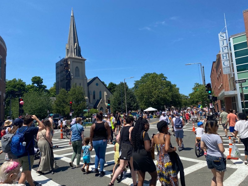 A large crowd of people walking and biking fills an intersection in front of a large church steeple.