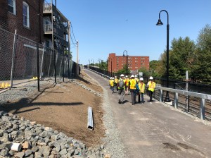 People wearing day-glo vests walk along a paved path that's under construction, with bare soil and riprap on the ground to the left and the Green Line Extension tracks below them to the right.