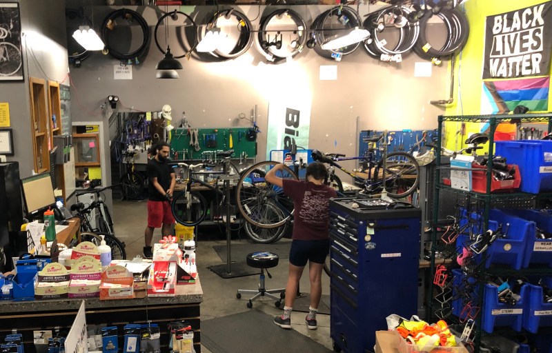 Two mechanics workon bikes on stands in a crowded workshop with bike tires hanging from the high ceilings.