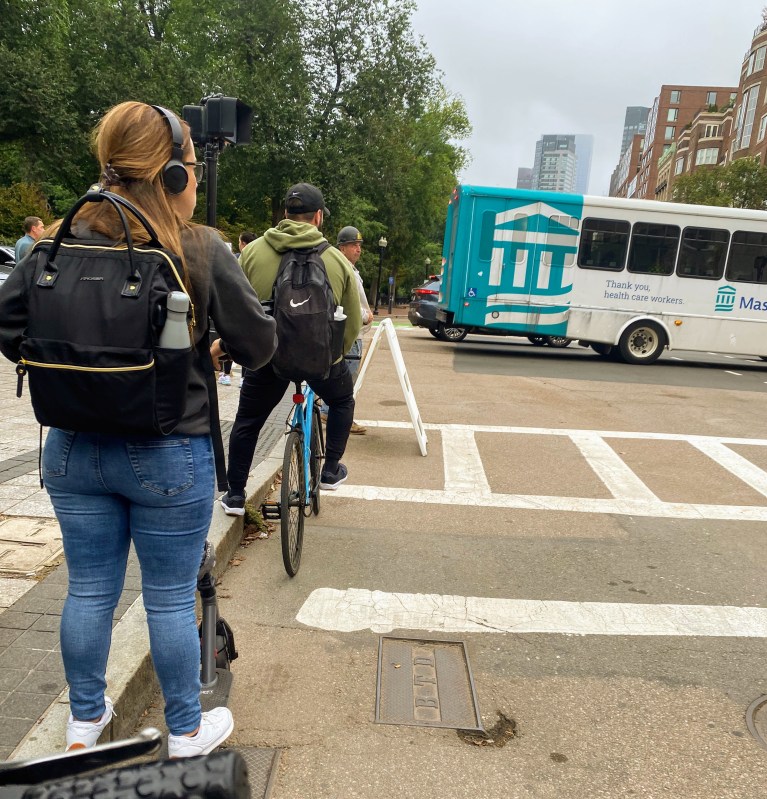 People on bikes and scooters wait at the intersection of Boylston Street and Arlington Street near the Public Garden in a pop-up bike lane that was installed during the Orange Line shutdown. In the background is the Public Garden, where an existing separated bike lane connects to the downtown bike network.