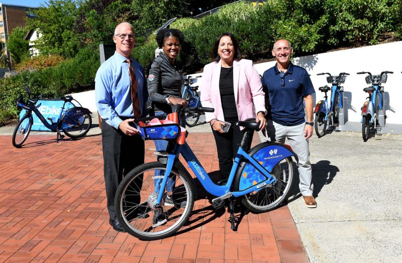 Four people: a white man wearing a blue collared shirt and tie, a black woman wearing an athletic long-sleeved top, a white woman wearing a pink blazer, and a white man wearing a collared short-sleeved polo shirt, stand behind a Bluebikes bicycle in a brick plaza. Behind them is a row of more bikes parked in a Bluebikes station.